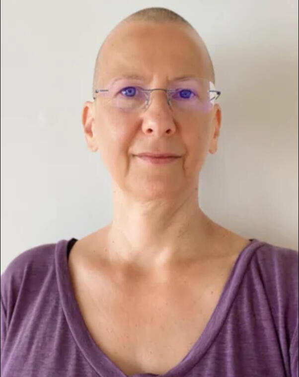 Daniela, a white woman with a shaved head, glasses, and a purple shirt, looks into the camera and smiles slightly.