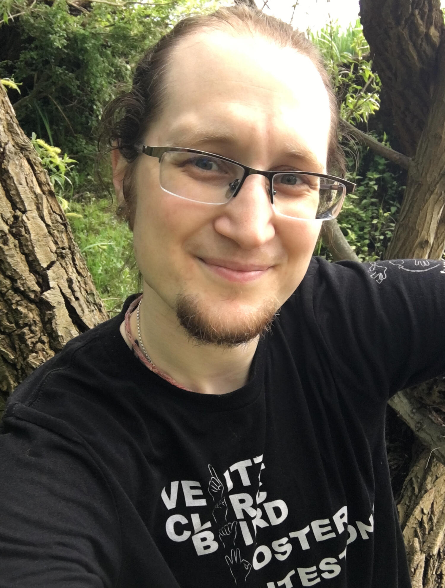 Rhys, a genderqueer person with glasses and short facial hair, looks into the camera and smiles.